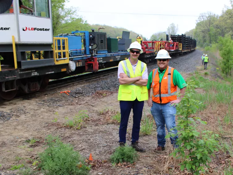 Two men smiling with hardhats and reflective vests standing in front of rail car loaded with 320' rail.