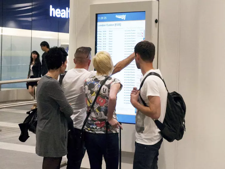 People using a wall mounted touchscreen to view journey times.