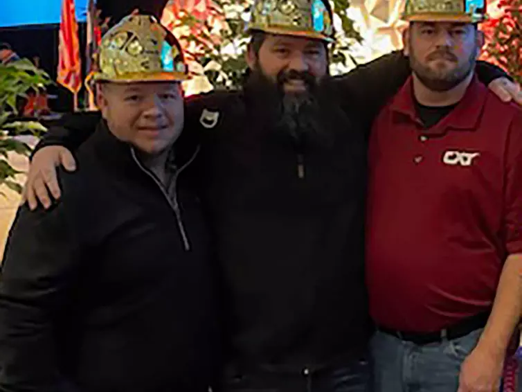 três homens com capacetes dourados sorrindo em frente a um palco no Precast Show.