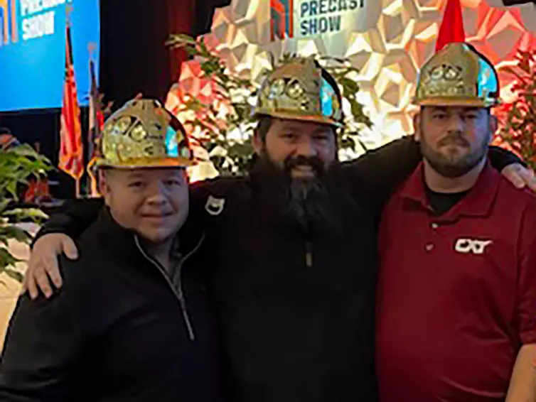 três homens com capacetes dourados sorrindo em frente a um palco no Precast Show.