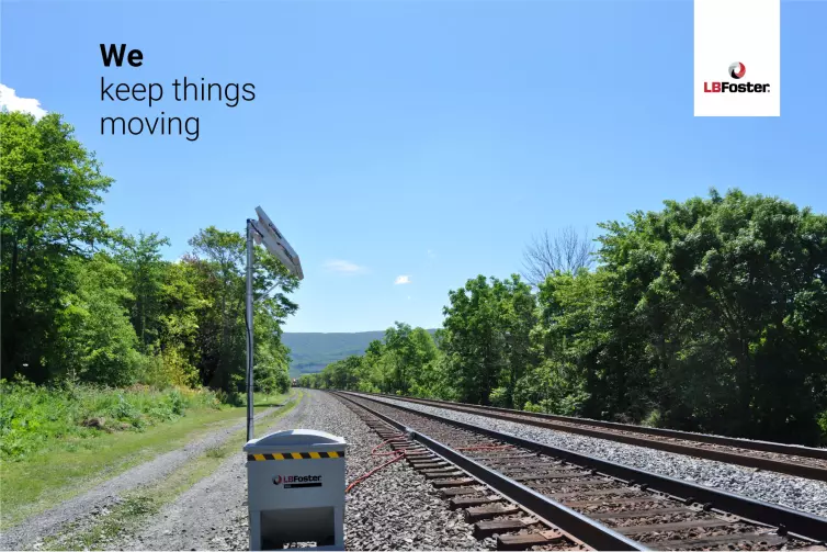 A protector unit and solar panel stands in the foreground with red tubing coming from it and out towards rail tracks. The tracks go far out into the distance and a train can be seen coming around the bend. There are trees either side of the tracks. Text on the image says 