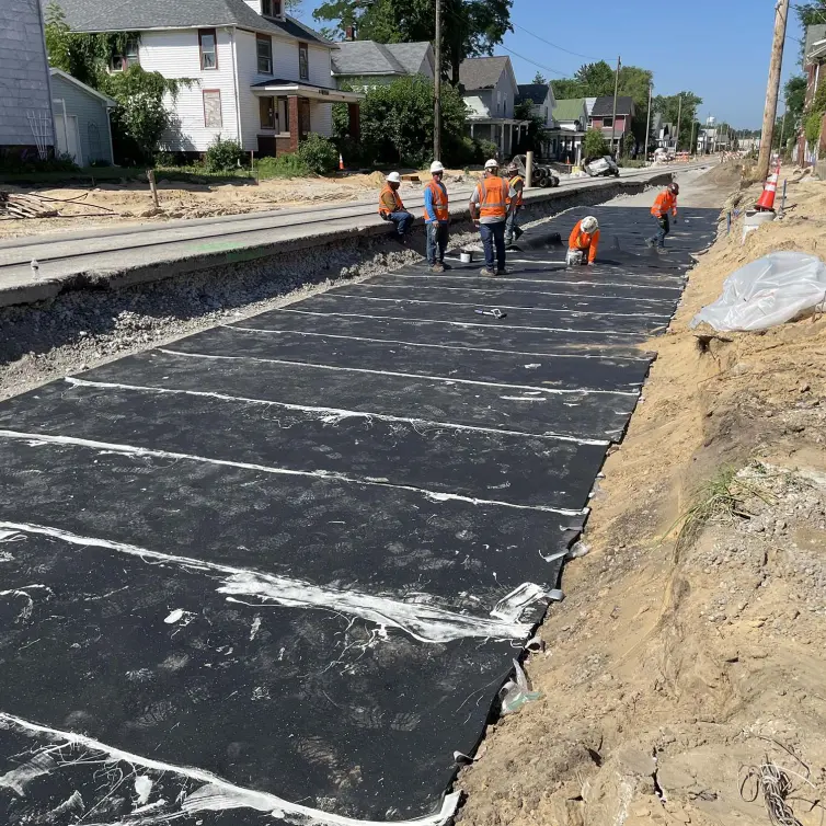 Six travailleurs portant des casques de sécurité et des gilets orange installant un tapis de ballast à côté d’une voie ferrée avec des maisons en arrière-plan.