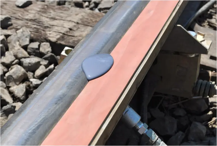 Primer plano de una vía férrea donde se ve un charco de líquido gris púrpura sobre una barra de espuma naranja sujeta al raíl.