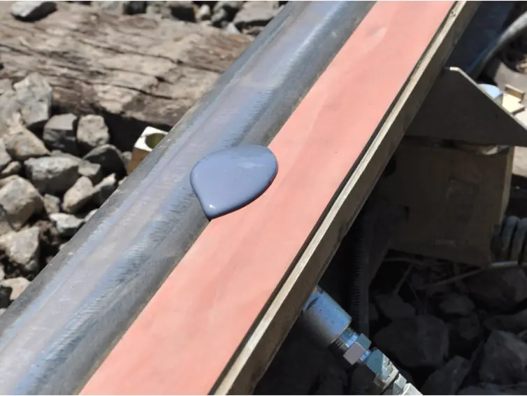 A close-up view of a railroad track where a puddle of purple-grey liquid is shown on an orange foam  bar attached to the rail.