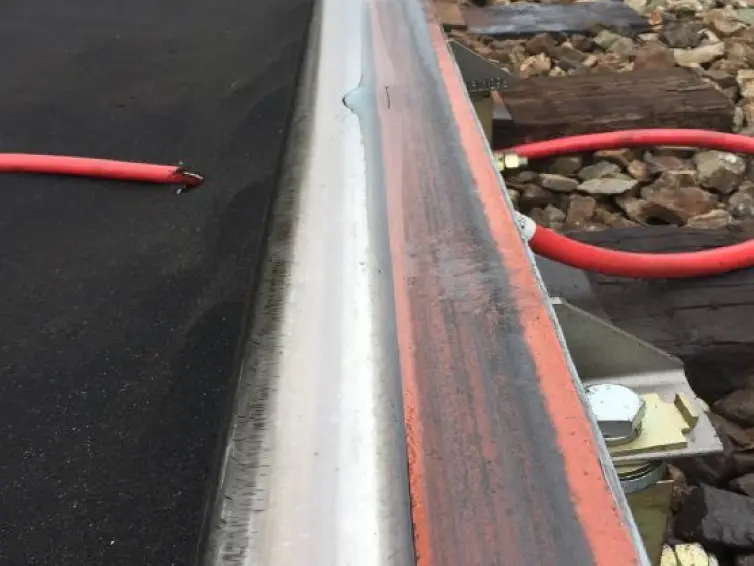 A close-up view of a railroad track where a puddle of clear  liquid is shown on an orange foam  bar attached to the rail.