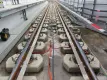 Ein Eisenbahngleis mit Betonplatten, auf denen die Schiene durch Befestigungselemente gestützt wird.  Stangen mit orangefarbenen Schläuchen sind an den Schienen befestigt, um das Produkt an der Oberseite der Schiene zu befestigen.