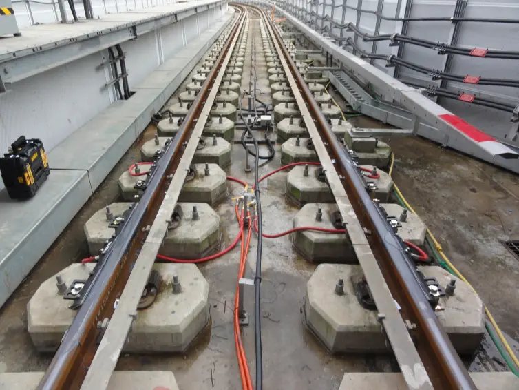 A railroad track with concrete slabs where the rail is supported by fasteners.  Bars with orange hoses are attached to the rails to product to the Top of the Rail.